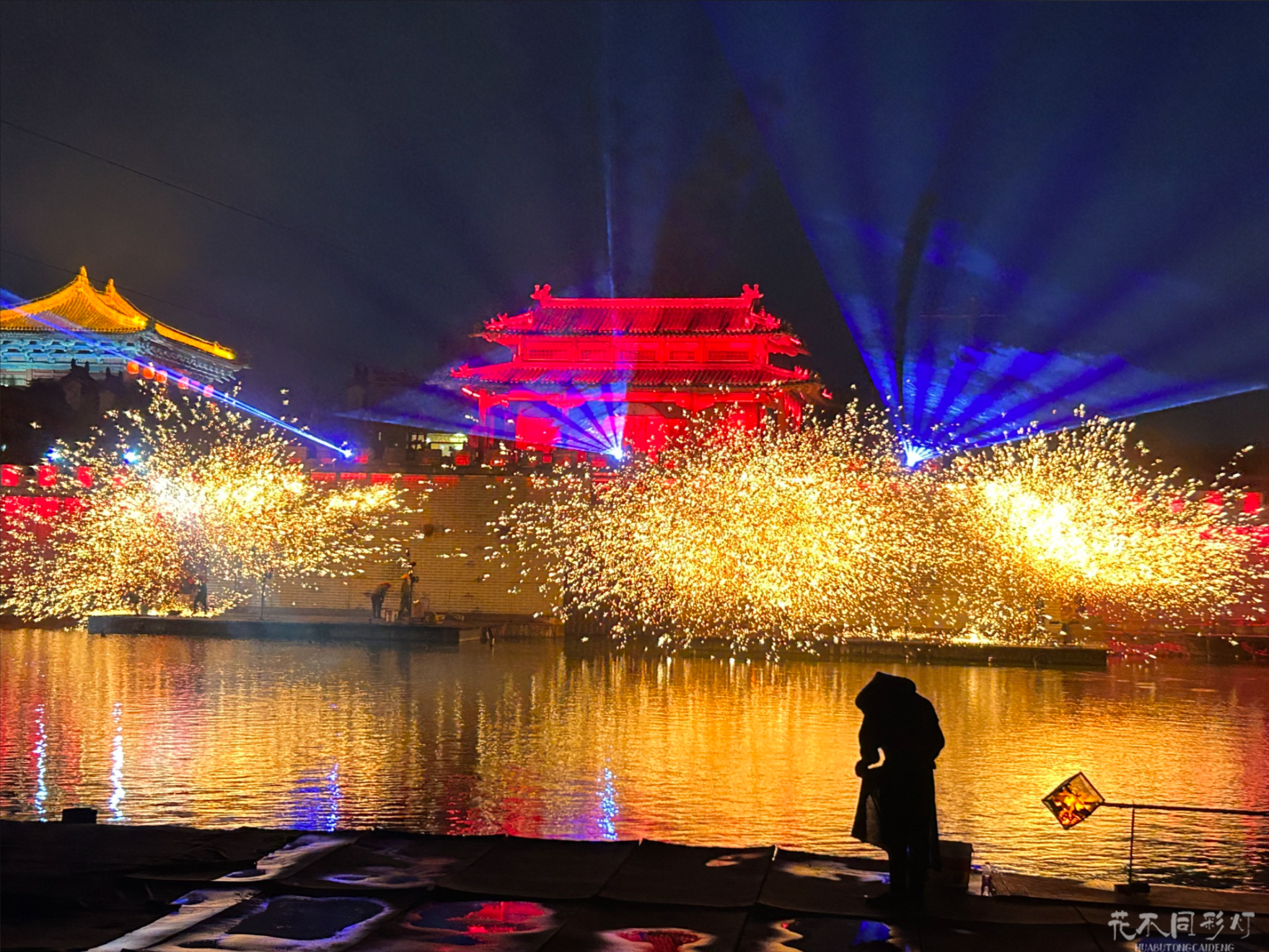 清明上河园夜景，璀璨的铁花在河面上空绽放，与水中的倒影形成对称的美景，游客在沿岸步道驻足观赏。